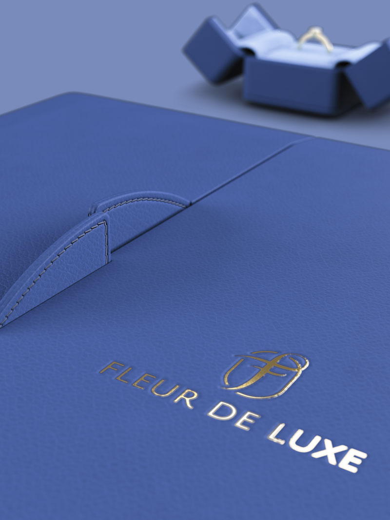 Détail de la couverture extérieure en cuir véritable d'un écrin à colliers Fleur de Luxe avec logo gaufré à la feuille d'or, derrière un écrin à anneaux ouvert